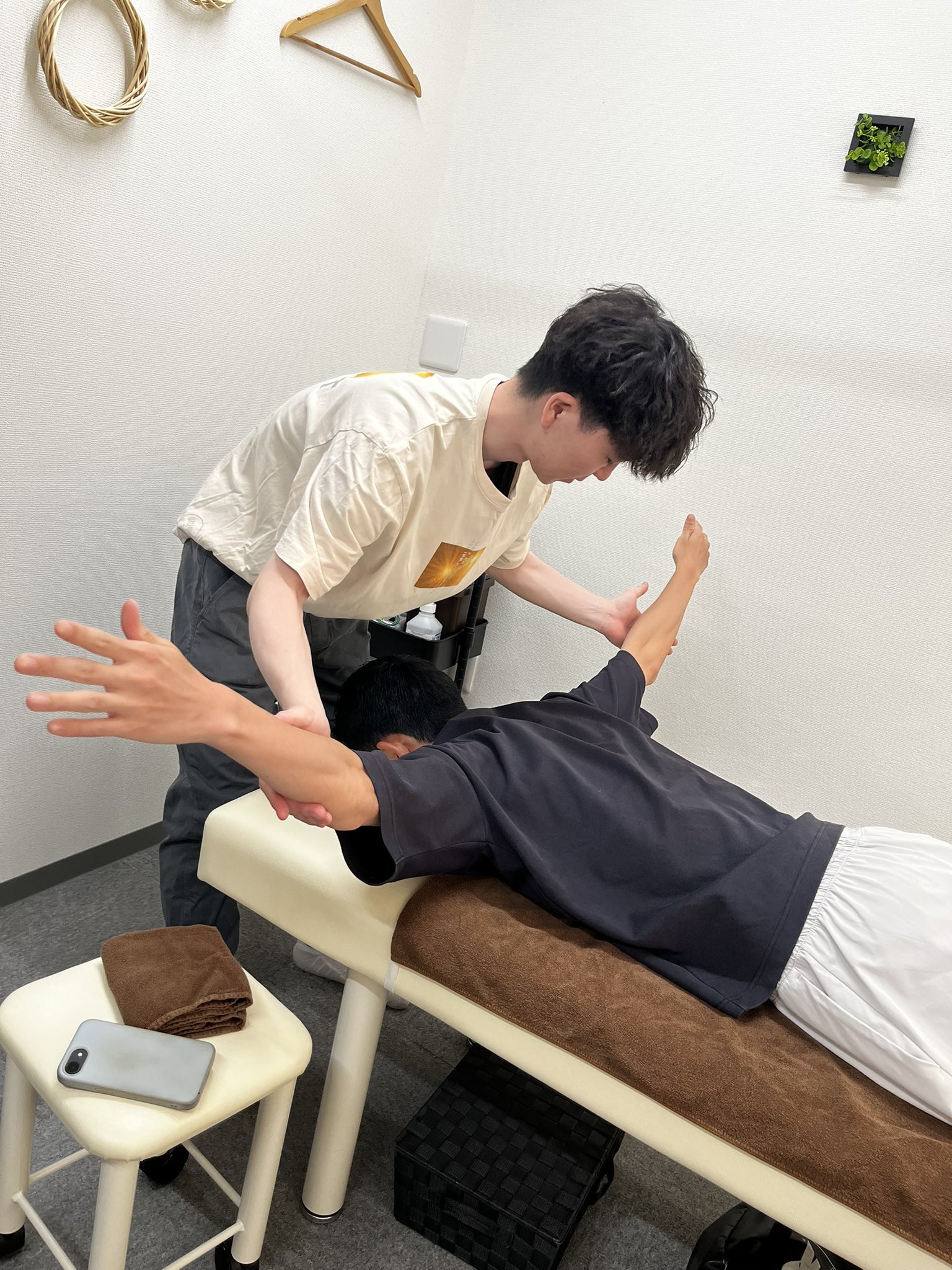頚肩部の運動療法の練習。うつ伏せで腕を挙げる種目を、肩甲骨に触れて誘導している場面