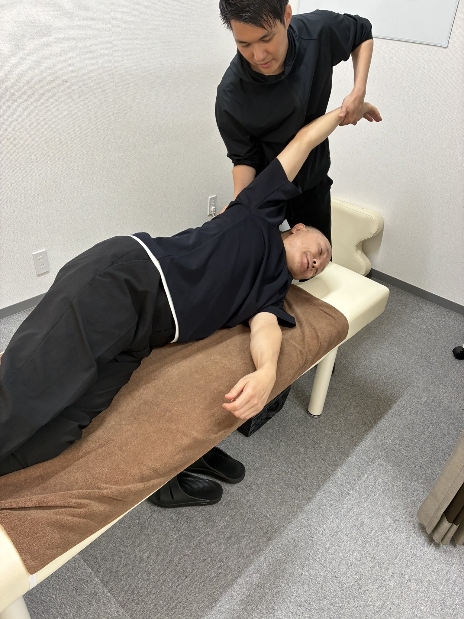 横向きの肢位で腕を天井方向へ挙げる種目を、施術者が支えて誘導している練習場面