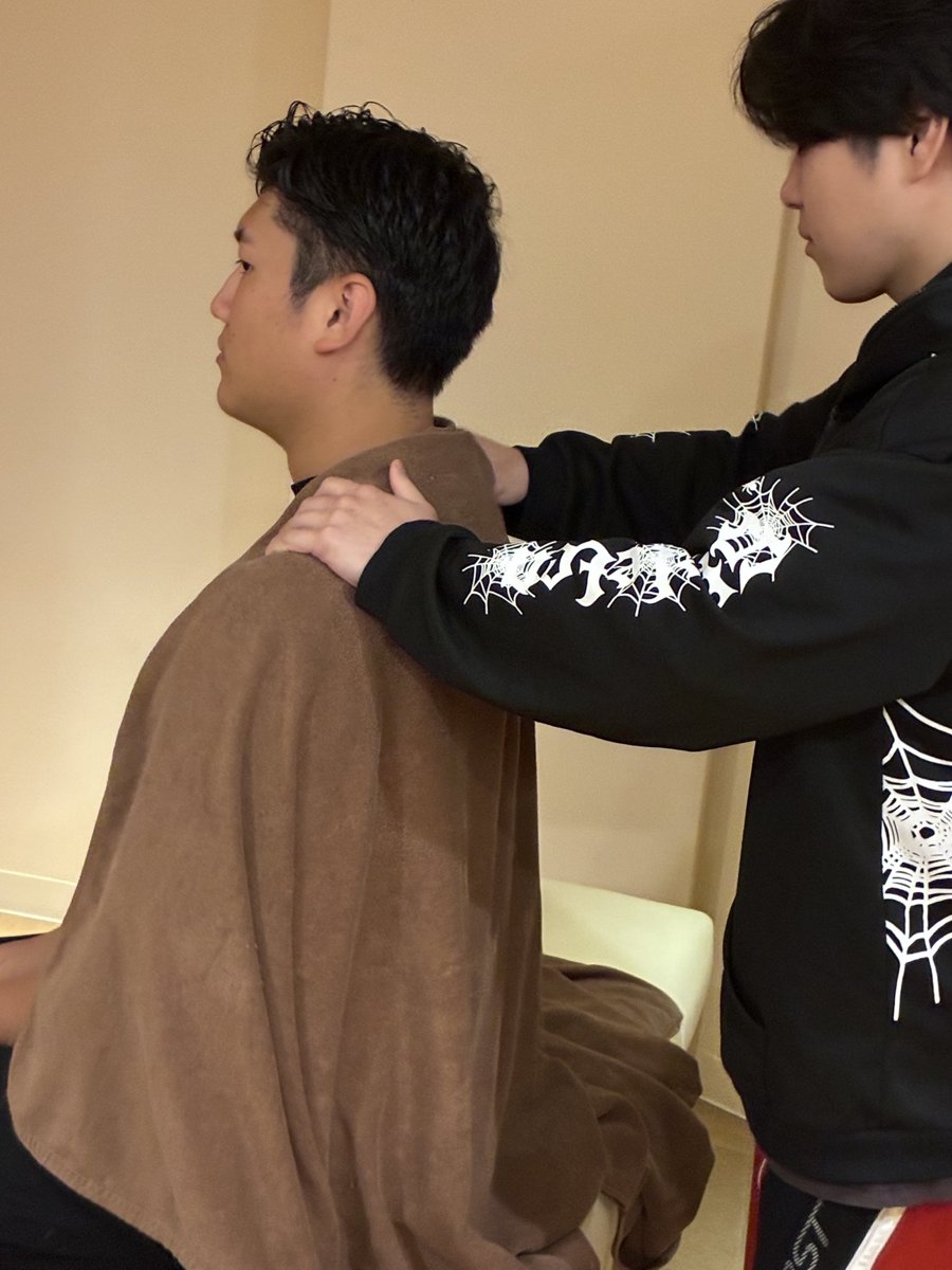 とんとん整骨院の練習会で、椅子に座ったスタッフの両肩に後ろから手を当てて肩まわりの施術を練習するスタッフ
