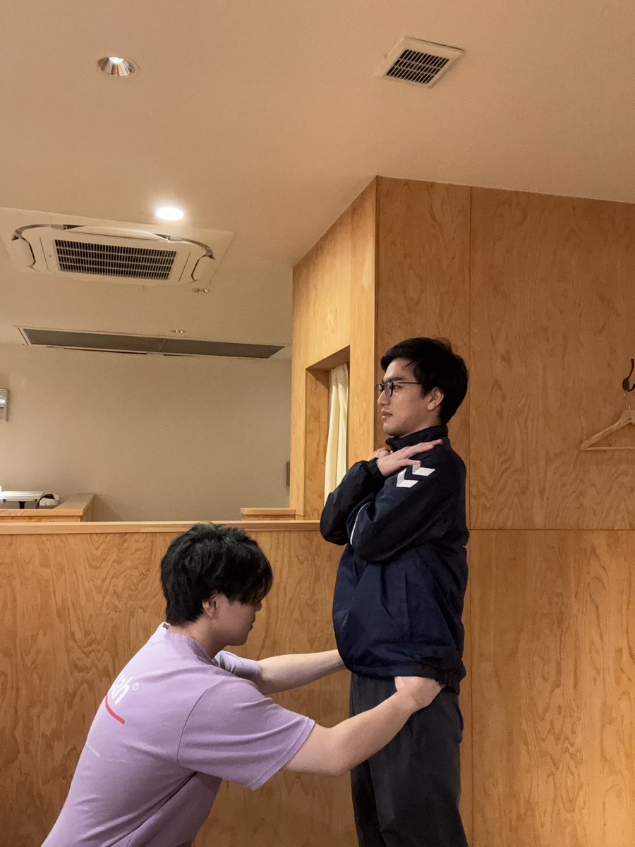 とんとん整骨院の練習会で、ベッドに仰向けになった受け役のスタッフの頭側に座り、首まわりに手を当てて施術の手順を確かめるスタッフ