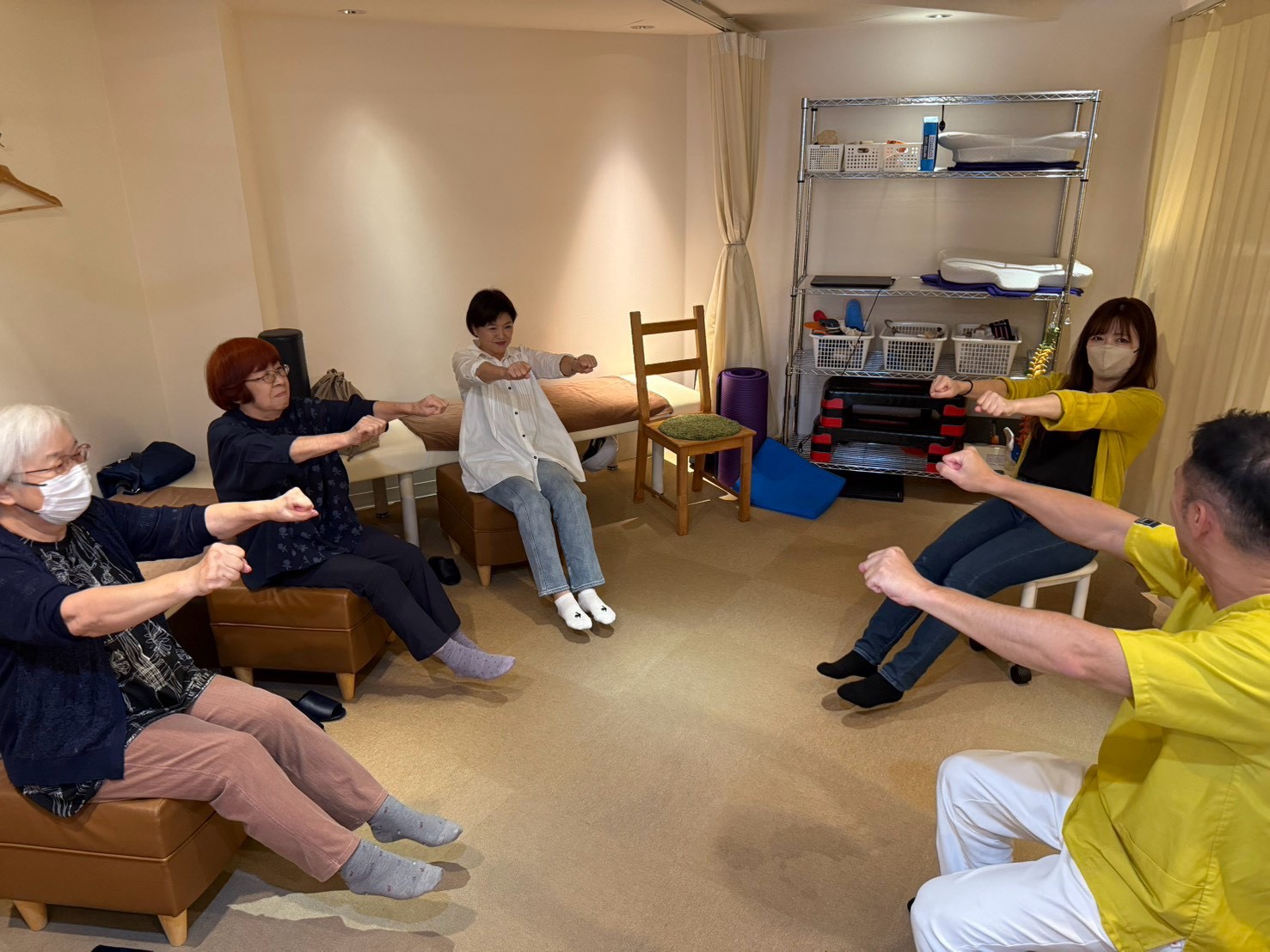 体操教室で、両手をグーにして前に出し、両足もそろえて前に伸ばす参加者とスタッフ