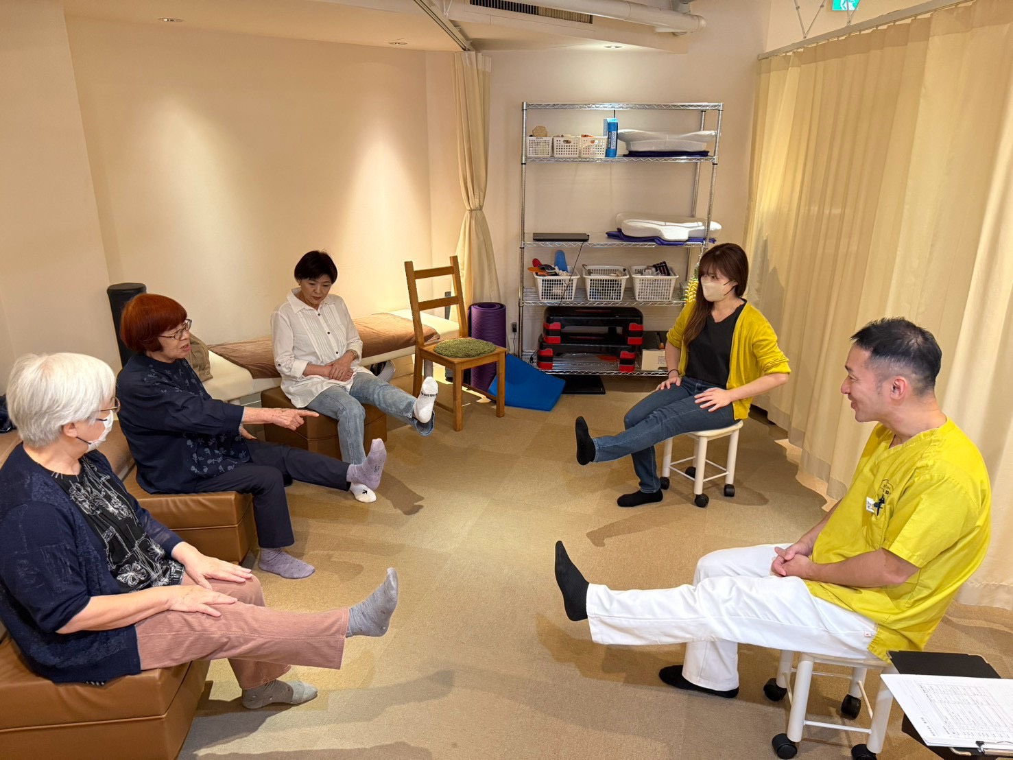 体操教室で、椅子に座ったまま片脚を前に上げる参加者とスタッフ